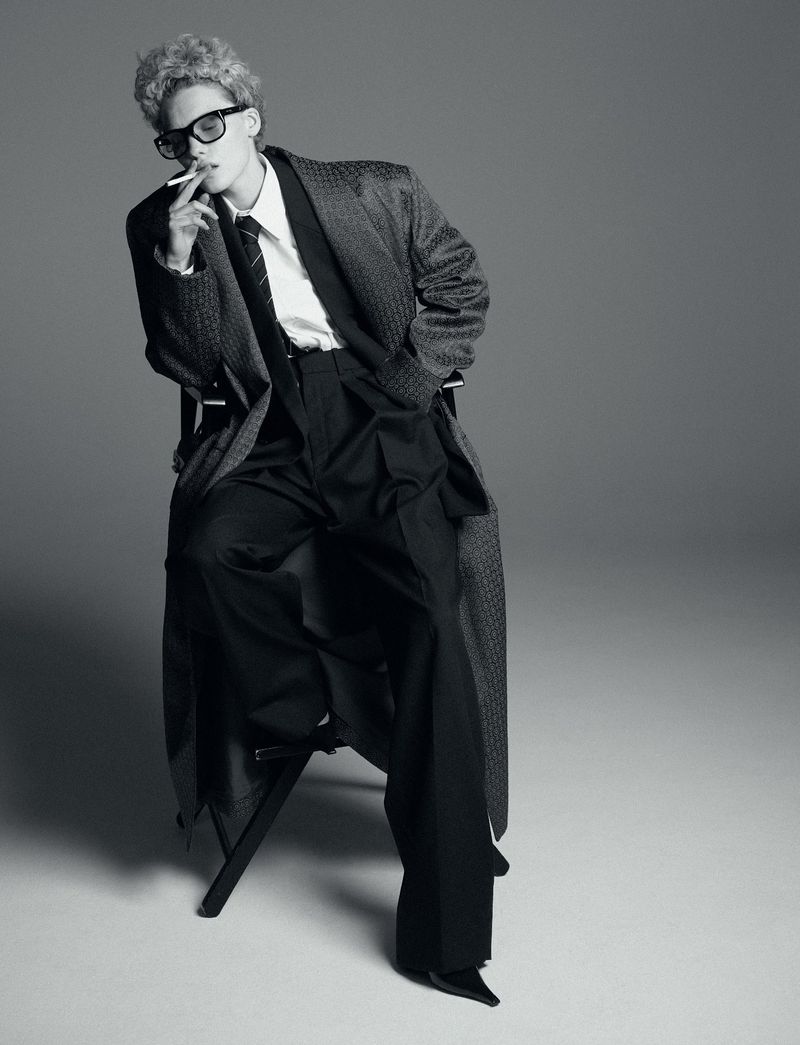 Black and white portrait of a person with curly hair, wearing a coat, shirt, and sunglasses, while seated and smoking a cigarette.