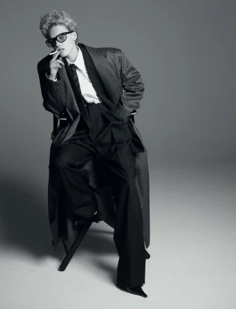 Black and white portrait of a person with curly hair, wearing a coat, shirt, and sunglasses, while seated and smoking a cigarette.