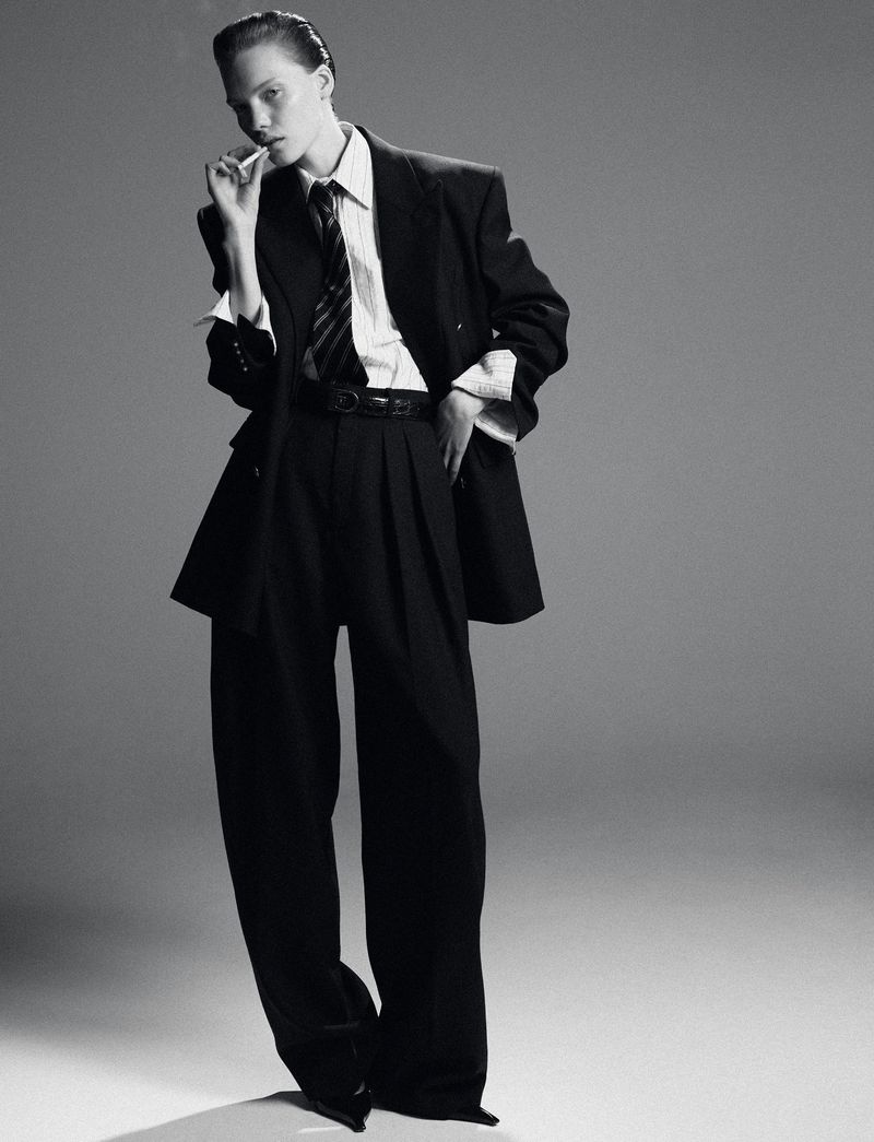 A woman in a black suit and tie strikes a confident pose against a grey background in this striking studio shot, embodying modern fashion.