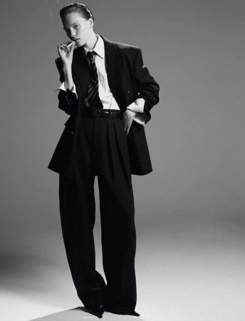 A woman in a black suit and tie strikes a confident pose against a grey background in this striking studio shot, embodying modern fashion.