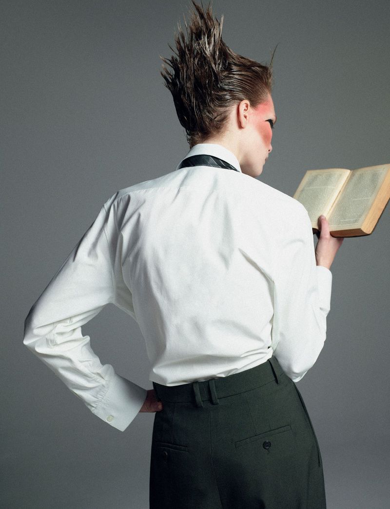 A person with a spiky hairstyle, seen from the back, is reading a book while wearing a white shirt and dark green pants in a studio.
