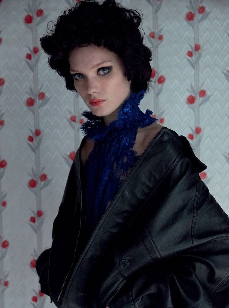 A model with dark curly hair and a blue lace collar poses in front of a floral wallpaper, wearing a black leather jacket.