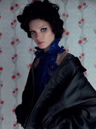 A model with dark curly hair and a blue lace collar poses in front of a floral wallpaper, wearing a black leather jacket.