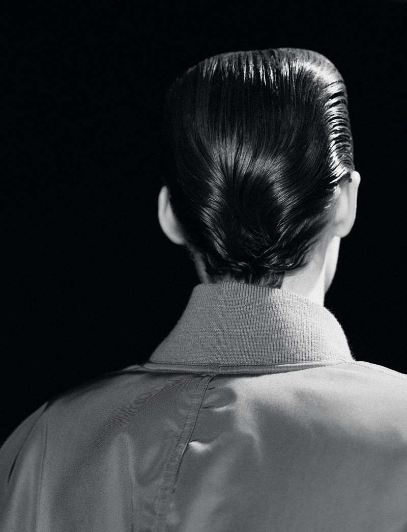 From behind, a person with a shiny pompadour hairstyle wears a light-coloured jacket against a stark black backdrop in a studio.
