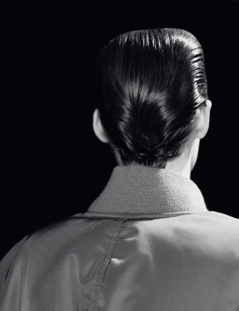 From behind, a person with a shiny pompadour hairstyle wears a light-coloured jacket against a stark black backdrop in a studio.