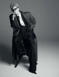 Model seated on a chair wearing oversized houndstooth coat, black trousers, white shirt, tie and dark glasses holding a cigarette