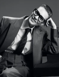 Model in oversized suit jacket and striped tie wearing thick black glasses against a grey background