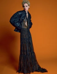 Model wearing a black leather jacket and long patterned dress posing against a solid orange background