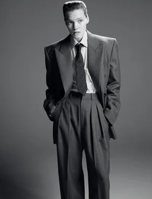 Model wearing an oversized grey suit jacket and trousers with a striped shirt and dark tie against a grey background