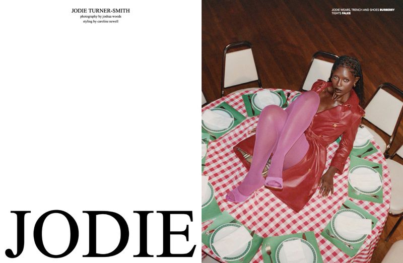 Editorial fashion shot of a Black female model wearing a red dress and pink tights, posing on a table with white plates and a checkered tablecloth.