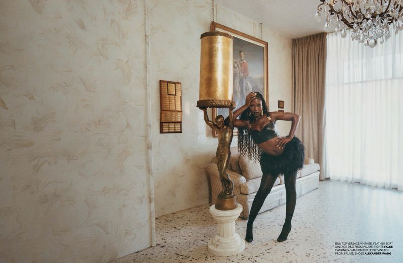 Elegant, dark-skinned woman strikes a pose indoors, sporting a black bra top and feathered skirt, beside a lamp featuring a gold statue.