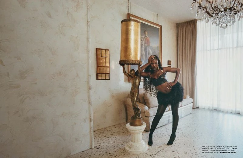Elegant, dark-skinned woman strikes a pose indoors, sporting a black bra top and feathered skirt, beside a lamp featuring a gold statue.