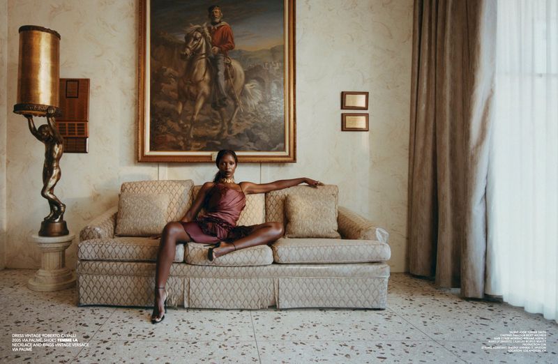 Elegant woman in vintage dress poses on sofa in ornate interior. The scene features an antique lamp, painting, neutral tones, and terrazzo floor.