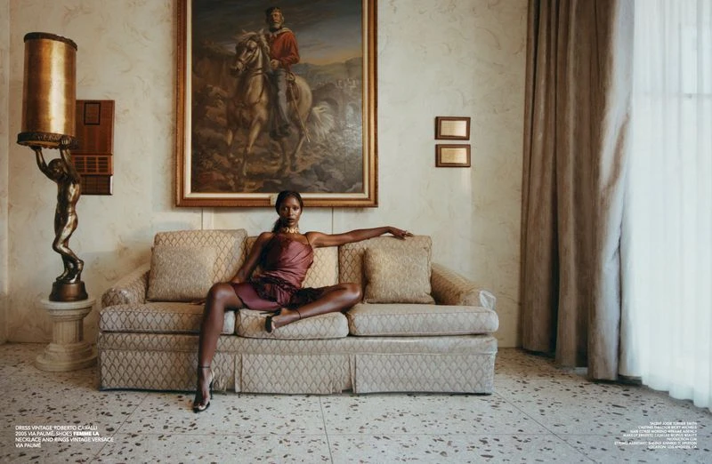 Elegant woman in vintage dress poses on sofa in ornate interior. The scene features an antique lamp, painting, neutral tones, and terrazzo floor.