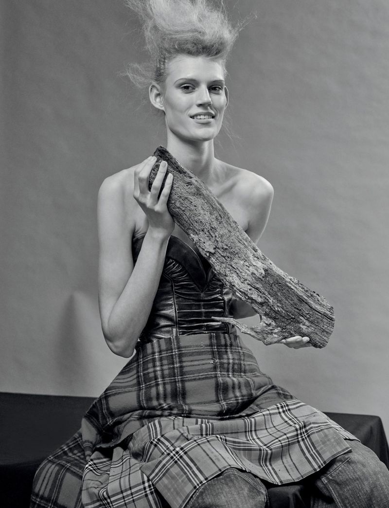 Monochrome fashion portrait of a woman with striking hair, holding a log, wearing a tartan skirt and a fitted top, against a plain backdrop.