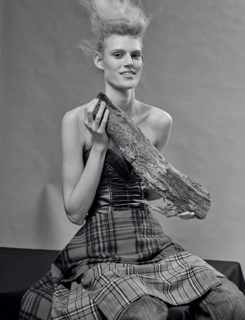 Monochrome fashion portrait of a woman with striking hair, holding a log, wearing a tartan skirt and a fitted top, against a plain backdrop.