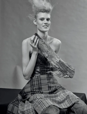 Monochrome fashion portrait of a woman with striking hair, holding a log, wearing a tartan skirt and a fitted top, against a plain backdrop.