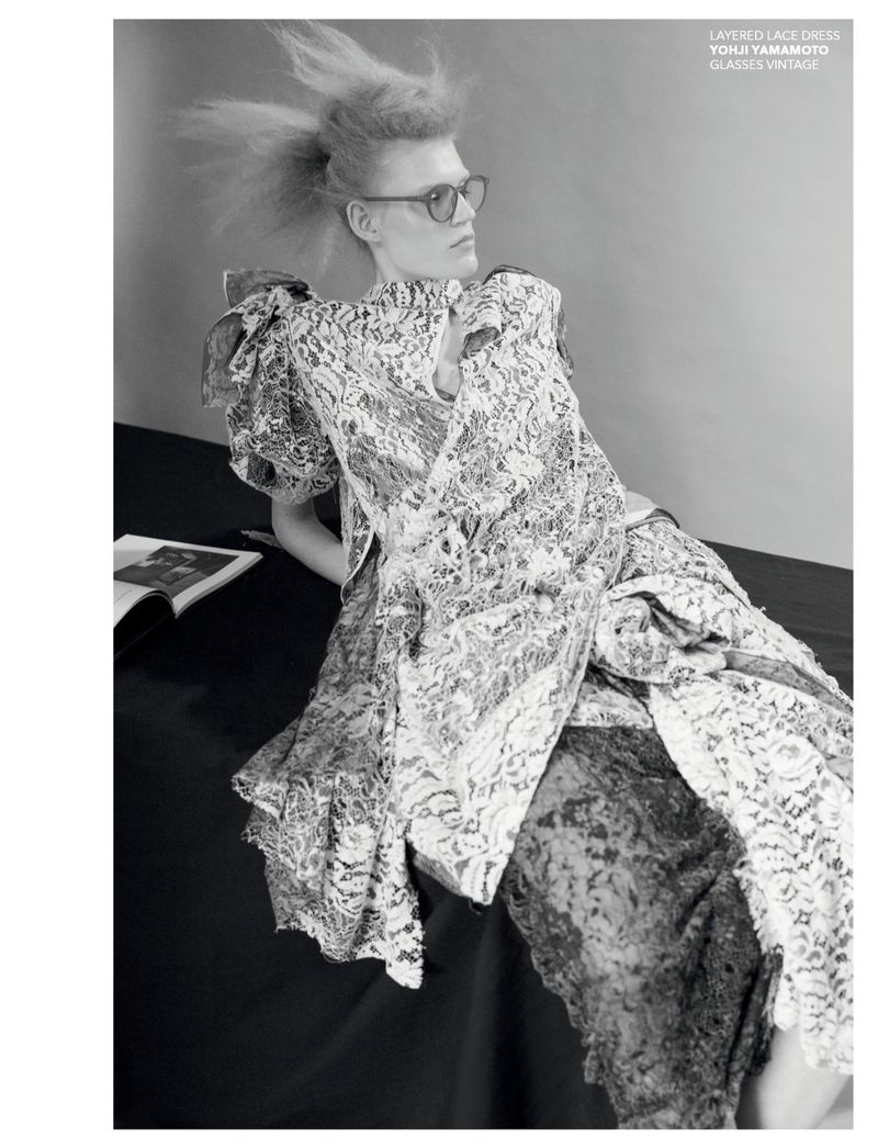 Monochrome fashion shot: a blonde model in a layered lace dress and glasses, lying on a dark surface with a magazine nearby.