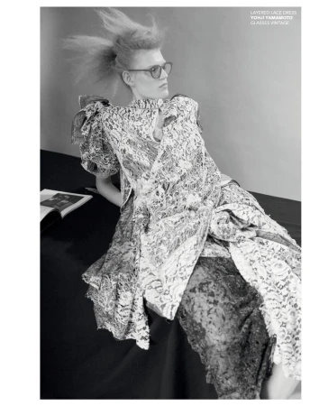 Monochrome fashion shot: a blonde model in a layered lace dress and glasses, lying on a dark surface with a magazine nearby.