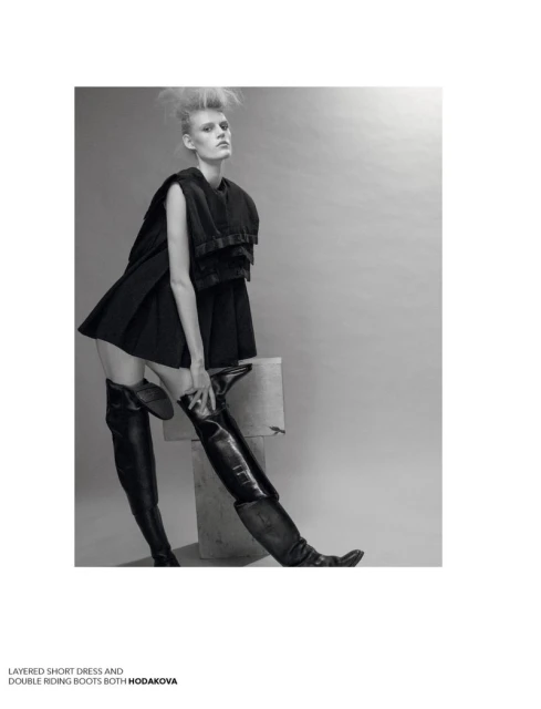 A fashion photo features a slender woman in a short black dress and thigh-high leather boots, posing against a neutral gray backdrop.