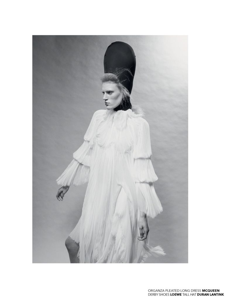 Monochrome fashion shot of a woman wearing a white pleated dress, derby shoes, and a tall black hat, set against a neutral background.