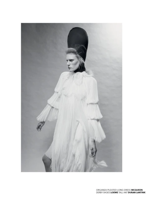 Monochrome fashion shot of a woman wearing a white pleated dress, derby shoes, and a tall black hat, set against a neutral background.