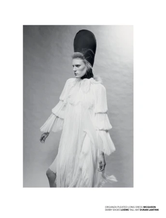 Monochrome fashion shot of a woman wearing a white pleated dress, derby shoes, and a tall black hat, set against a neutral background.