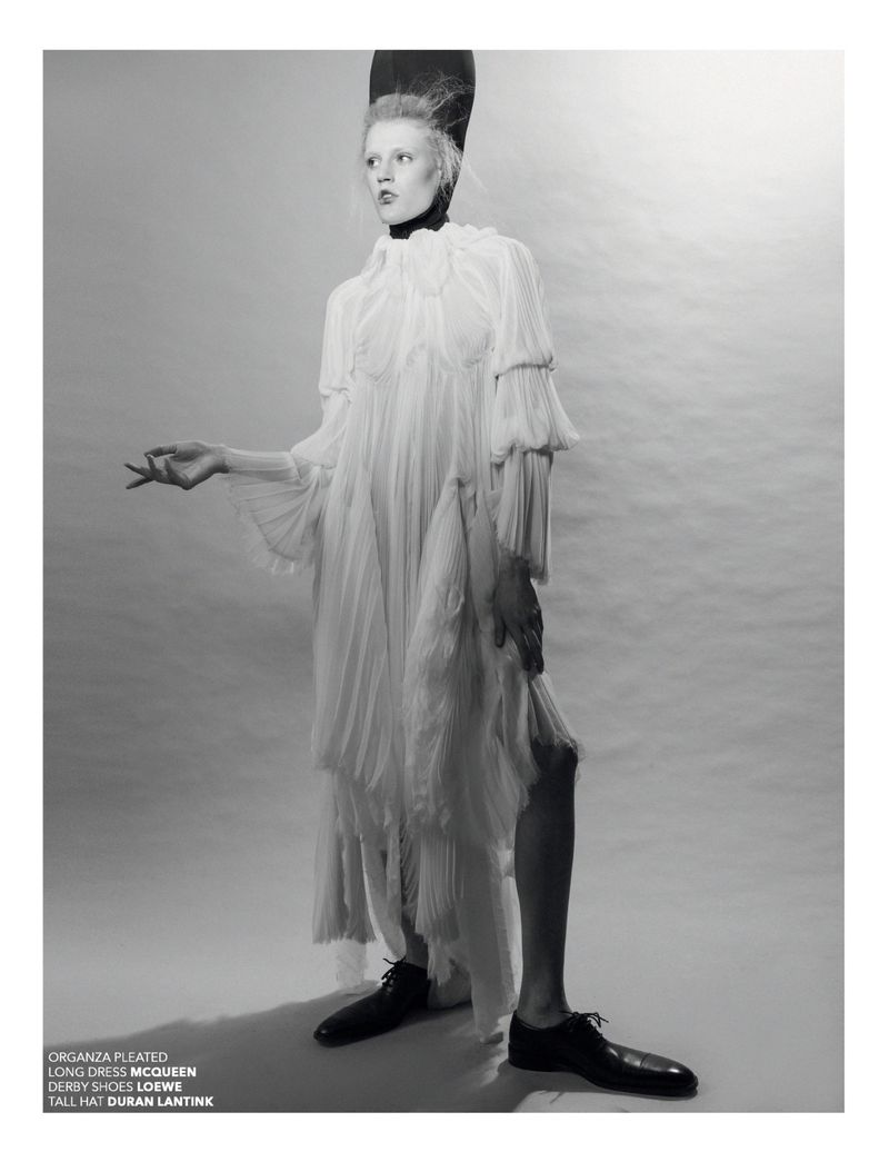 A model posing in a studio wearing a pleated long dress and a tall hat in a fashion editorial shot with monochromatic color grading.