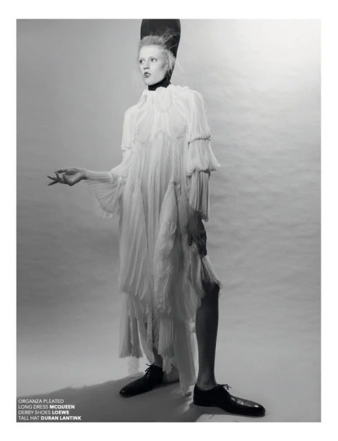 A model posing in a studio wearing a pleated long dress and a tall hat in a fashion editorial shot with monochromatic color grading.