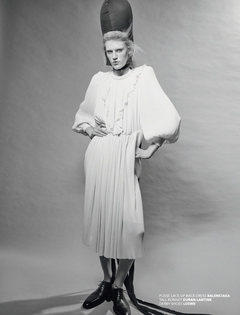 A model in a white dress and bonnet poses in a studio. The black and white image features fashion elements and studio lighting.