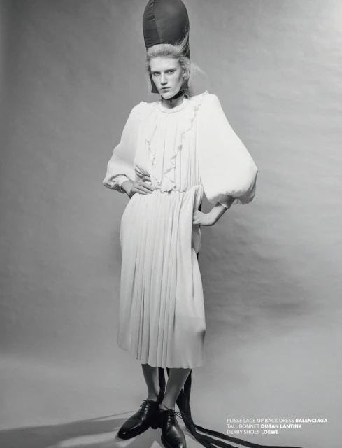 A model in a white dress and bonnet poses in a studio. The black and white image features fashion elements and studio lighting.