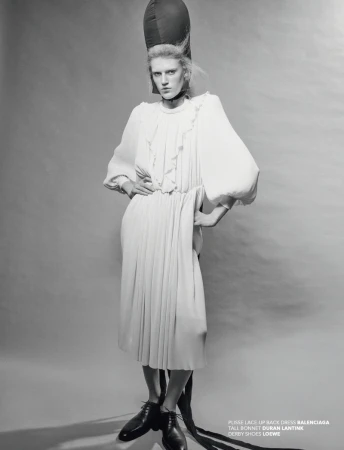 A model in a white dress and bonnet poses in a studio. The black and white image features fashion elements and studio lighting.