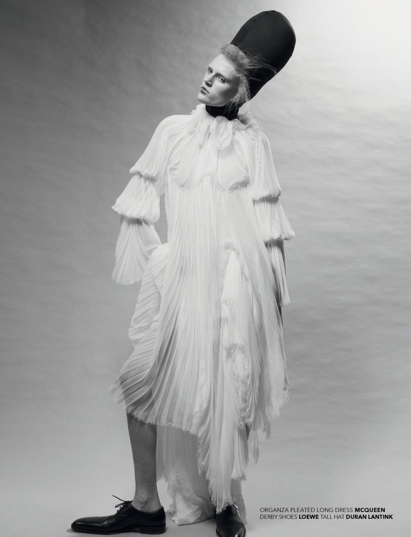 A full-length studio portrait of a slender adult fashion model wearing a long white pleated dress and a tall black hat against a grey backdrop.