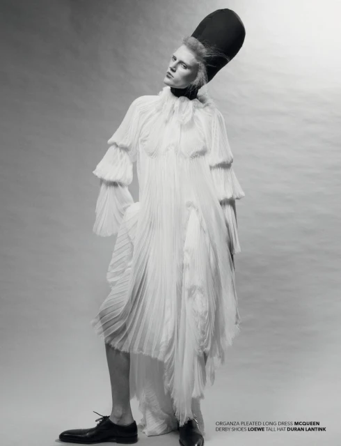 A full-length studio portrait of a slender adult fashion model wearing a long white pleated dress and a tall black hat against a grey backdrop.