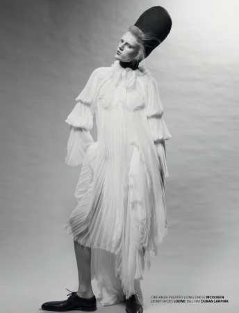 A full-length studio portrait of a slender adult fashion model wearing a long white pleated dress and a tall black hat against a grey backdrop.