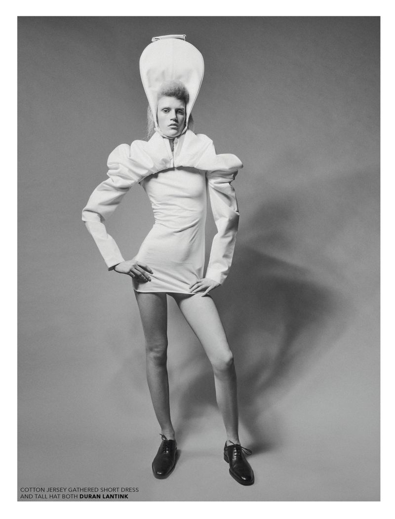 Black and white fashion shot of a model in a short white dress with puffy sleeves and a tall white hat, posing with hands on hips.