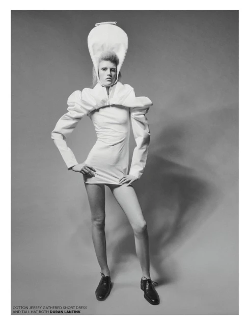 Black and white fashion shot of a model in a short white dress with puffy sleeves and a tall white hat, posing with hands on hips.