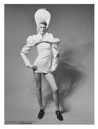 Black and white fashion shot of a model in a short white dress with puffy sleeves and a tall white hat, posing with hands on hips.