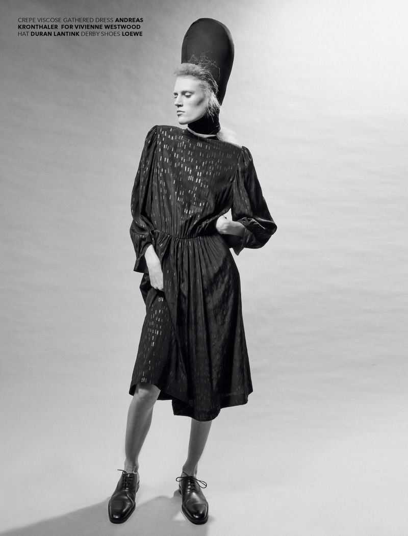 A slender model poses in a patterned midi dress with a tall hat. She wears derby shoes in a studio setting with diffused lighting.