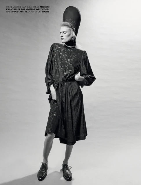 A slender model poses in a patterned midi dress with a tall hat. She wears derby shoes in a studio setting with diffused lighting.