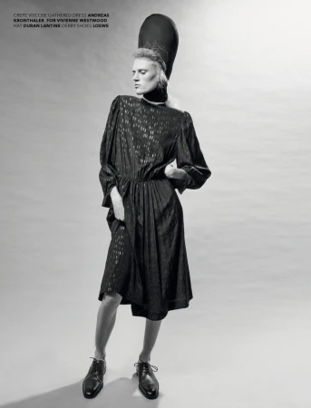 A slender model poses in a patterned midi dress with a tall hat. She wears derby shoes in a studio setting with diffused lighting.