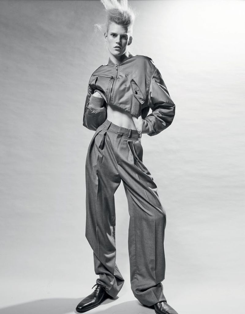 Monochrome studio shot of a young adult fashion model with unique hairstyle wearing a cropped jacket and wide-leg pants, hands on hips.
