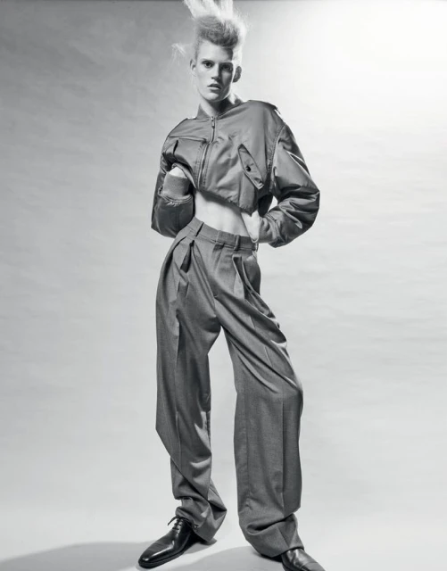 Monochrome studio shot of a young adult fashion model with unique hairstyle wearing a cropped jacket and wide-leg pants, hands on hips.