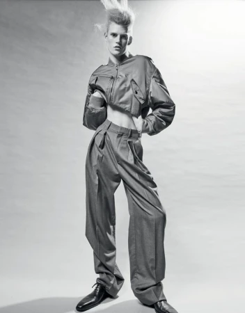 Monochrome studio shot of a young adult fashion model with unique hairstyle wearing a cropped jacket and wide-leg pants, hands on hips.