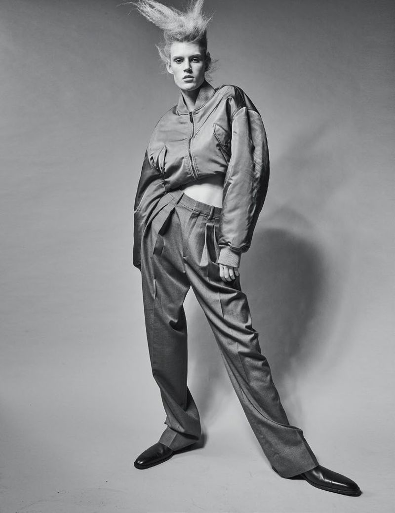 Monochrome fashion shot of a slender adult woman in a bomber jacket and wide-legged trousers, standing against a light grey backdrop.