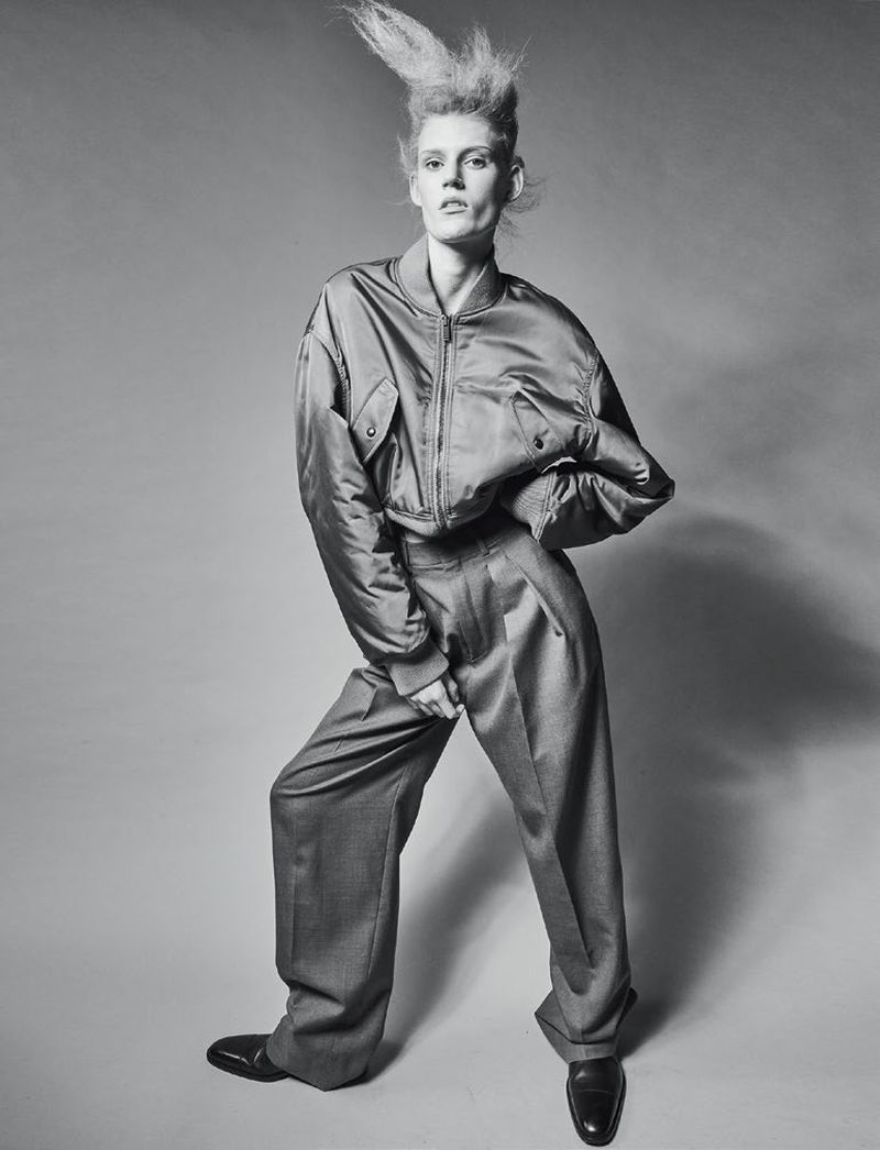 Fashion model poses in studio wearing an oversized satin bomber jacket and wide-legged wool trousers with glossy black shoes and spiked hair.