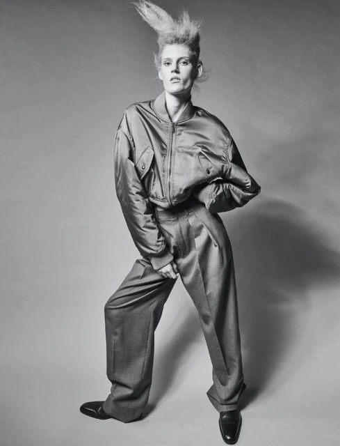 Fashion model poses in studio wearing an oversized satin bomber jacket and wide-legged wool trousers with glossy black shoes and spiked hair.