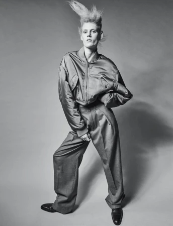 Fashion model poses in studio wearing an oversized satin bomber jacket and wide-legged wool trousers with glossy black shoes and spiked hair.