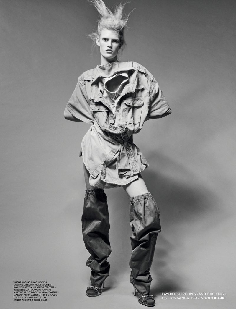 Fashion shot of a female model in a light-colored shirt dress and thigh-high boots standing against a grey studio backdrop with bright lighting.