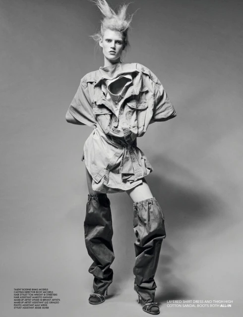 Fashion shot of a female model in a light-colored shirt dress and thigh-high boots standing against a grey studio backdrop with bright lighting.
